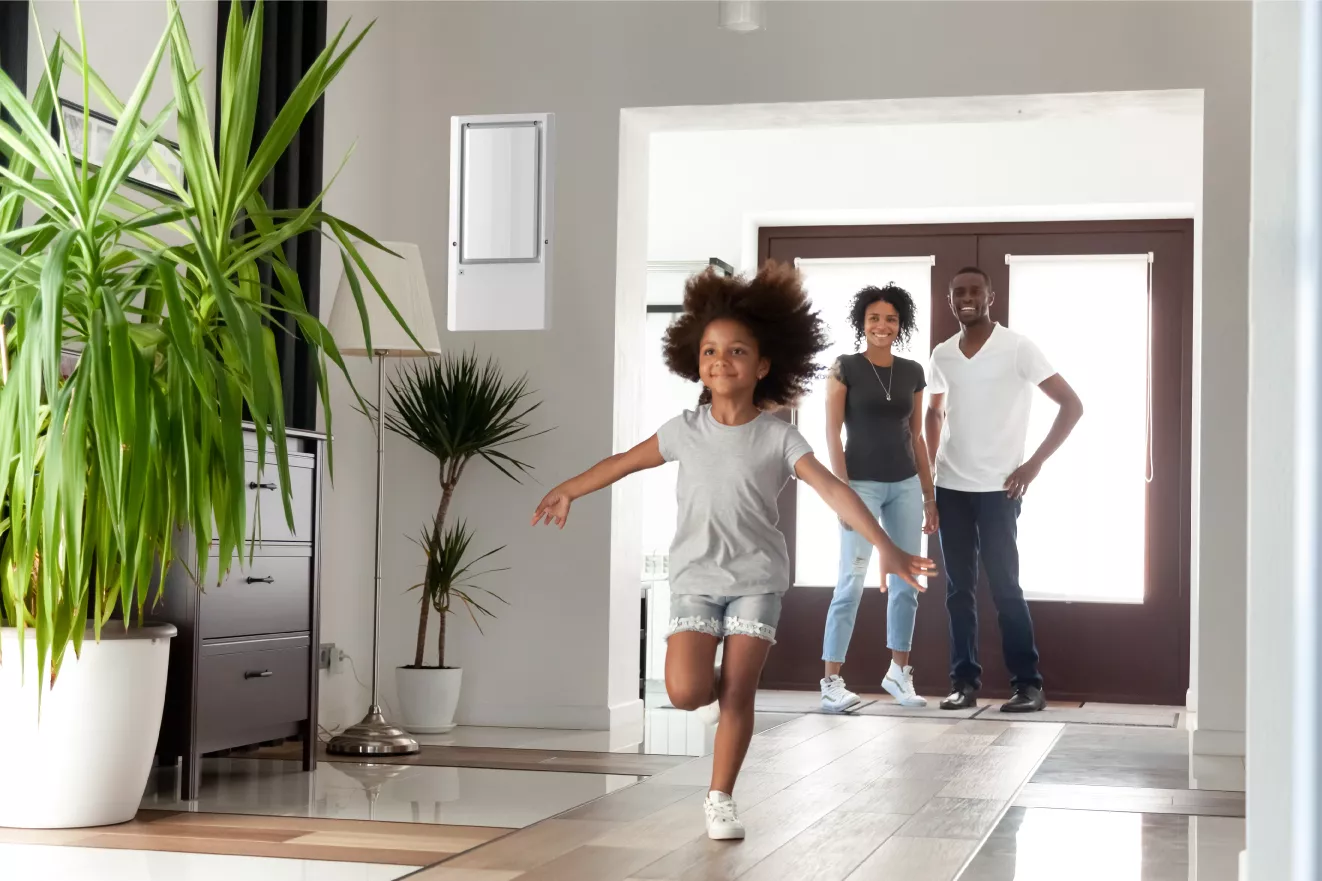 Young girl smiling as enters her home, parents watch with smiles. The IW25-4 fixed dehumidifier is installed on the back left wall above plants.